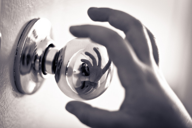 Black and white image of a hand reaching to grab a door handle. The door handle  has a reflection of this in action.