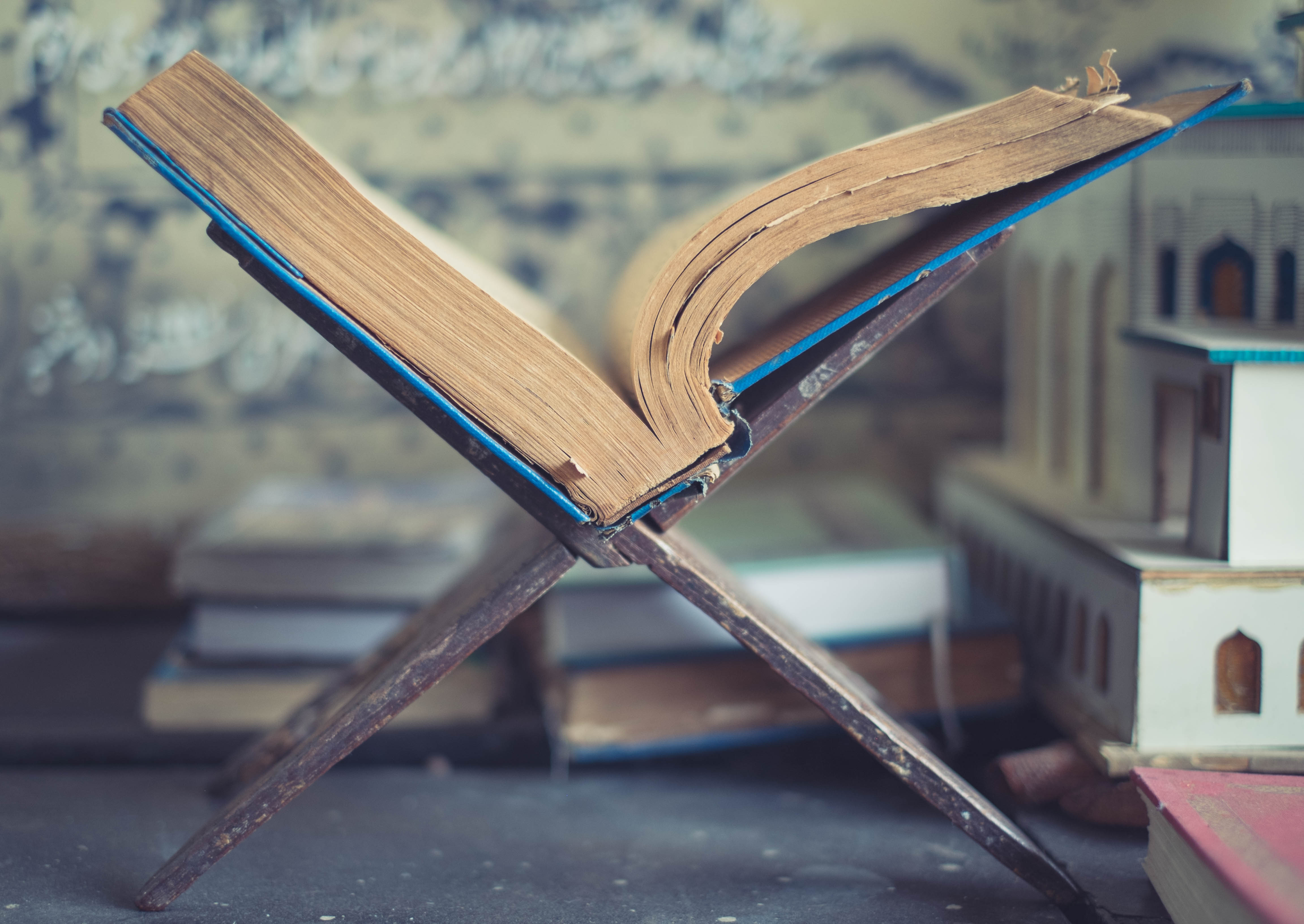 A book lies open in the middle, propped up by a book stand. Out of focus, in the background are several other books and religious artifacts.