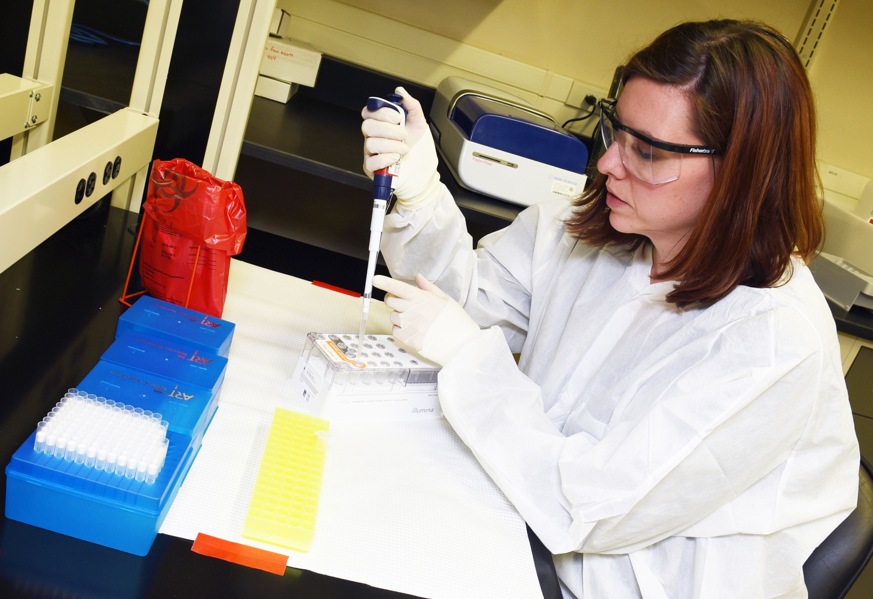 A laboratory scientist, in goggles, and protective gear, holds a pipette and concentrates on placing a sample of liquid into a test tube.