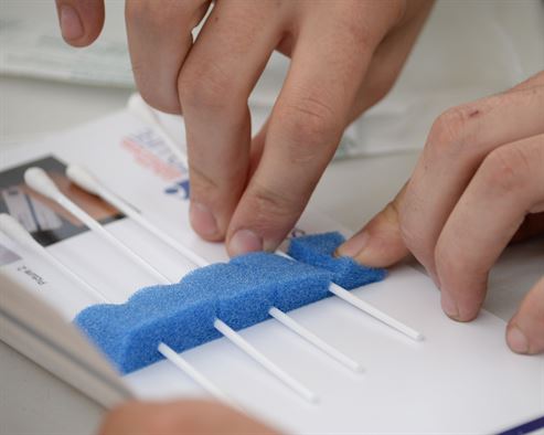 A mail-in kit, containing four long cotton swabs, placed in an envelope, with place holders for each cotton swab.