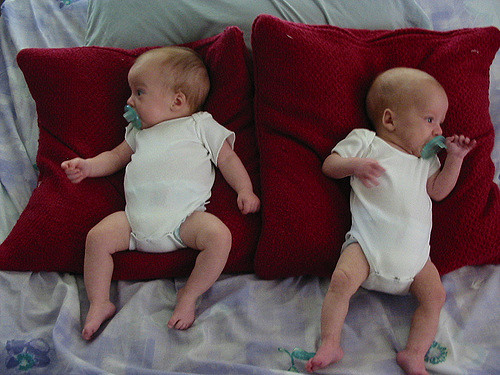 Two babies lay side by side on two separate pillows, appearing to sleep.