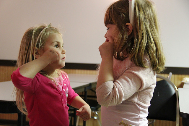 Two young children mirror each other in communicating through American Sign Language (possibly signing gum or candy).