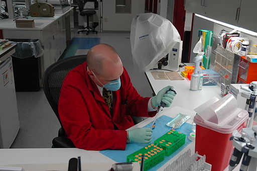 A male lab technician in a red coat and a blue face mask sits at a desk. He is concentrated on inserting a sample of DNA from a pipette into a test tube. A test tube rack is placed on his desk along with a hazardous waste basket and other equipment.