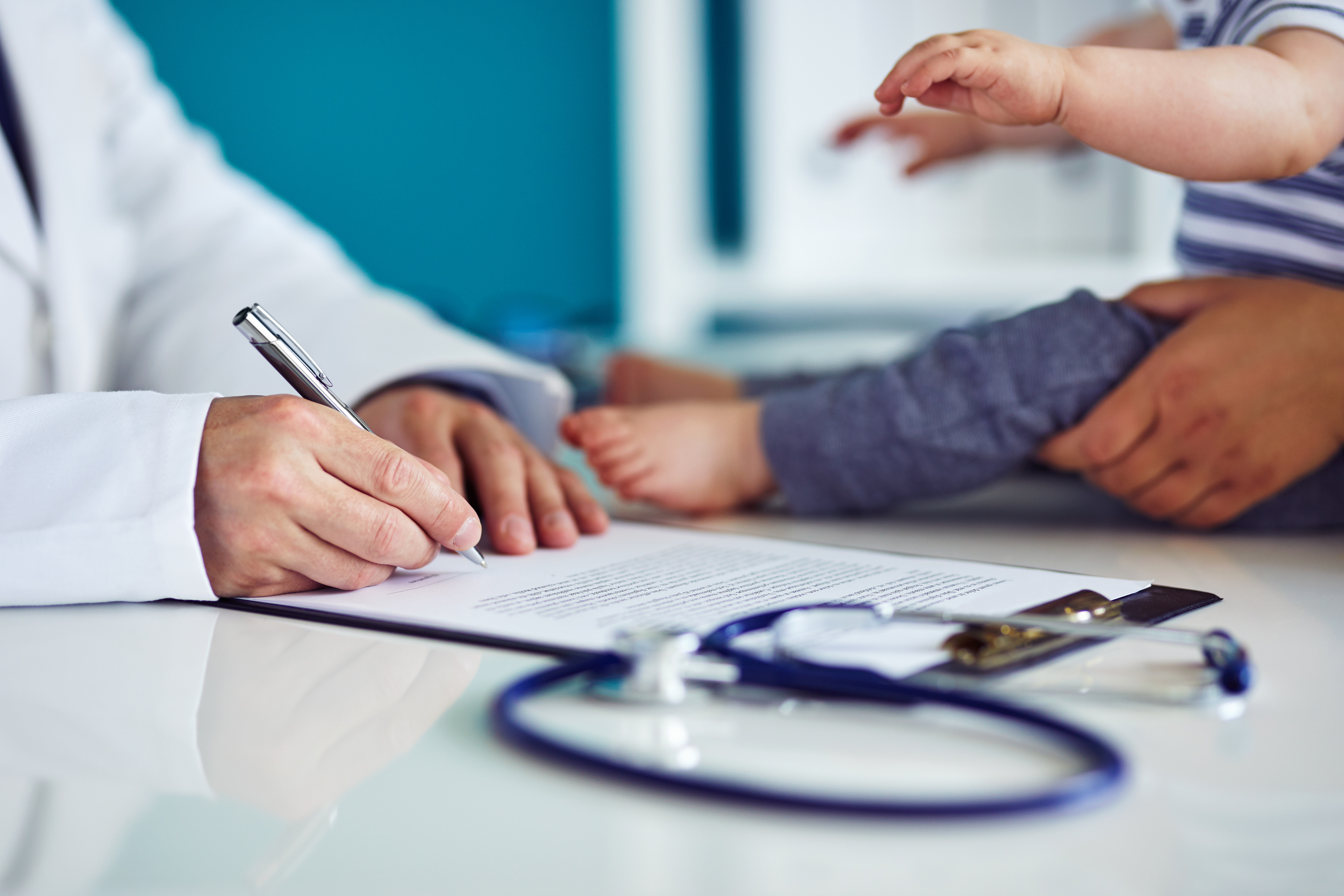 Doctor in white coat writing on a clipboard on a table next to an infant dressed in blue and a stethoscope