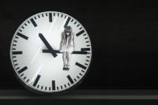 Black and white image. A woman sits peering down, on the long hand of a clock. The clock reads 10:15. 