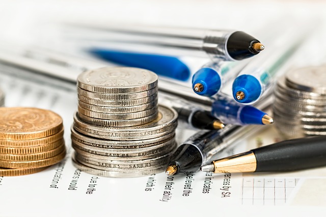 Three stacks of coins are resting on top of paper that has small font. Blue and black pens are stacked between the coin piles.