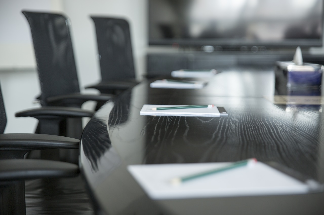 Zoomed into the edge of a brown conference table. Black chairs flank the table and notepads and pencils lie on top.