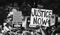 A handwritten sign held overhead at a protest reads "JUSTICE NOW" in all caps