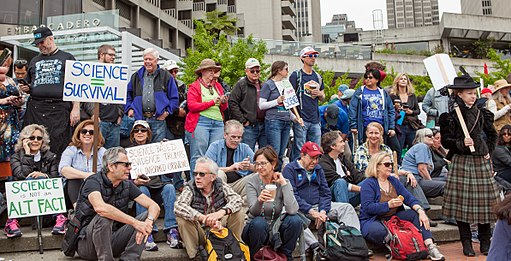 March for Science protesters sit and stand on staircase, and hold various signs and banners. In the background is the San Francisco Embarcadero.