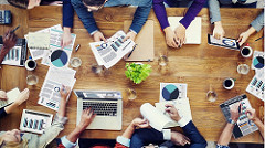 Bird's eye view of a large group of people who are seated around a round table, looking at laptops, and infographics, with coffee mugs placed on the table.