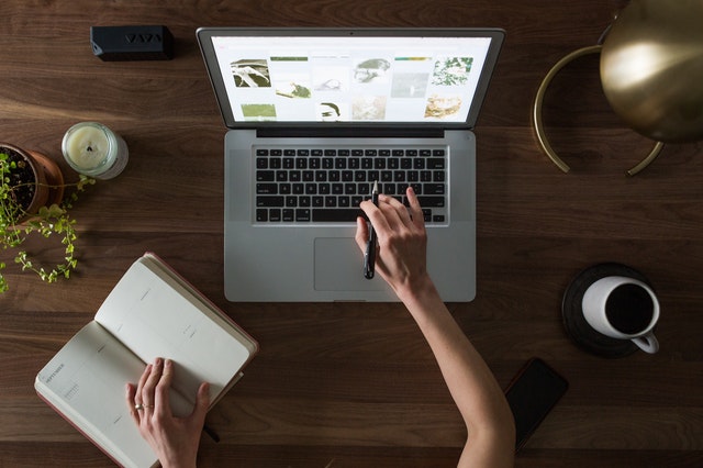 Bird's eye view of an adult with slender features working, using a laptop, and notebook and surrounded by coffee, plant and candle on a wooden desk.