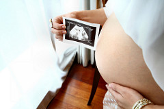 Bird's eye view of the side of a pale skinned pregnant woman, with an exposed stomach, who uses one hand to grasp her stomach, and the other to hold up a sonogram of the baby.