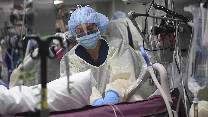 A nurse, fully covered in PPE, leans over a hospital bed surrounded by medical equipment