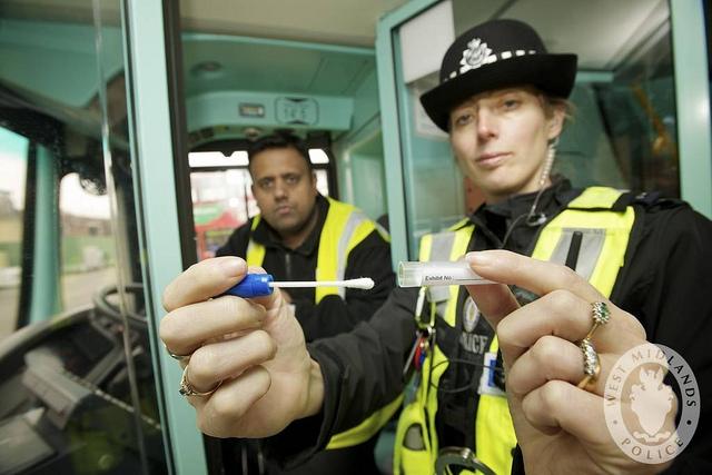 Two authorities in yellow jackets look directly at the camera, with an eye of suspicion. One of them holds out a DNA swabbing spit kit.