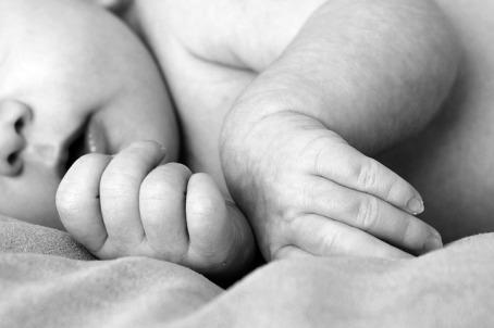 Black and white image of a baby laying down. The baby's hands and fingers appear gently curled and are the focal point of the picture. In the background, the baby's nose and mouth are featured. 