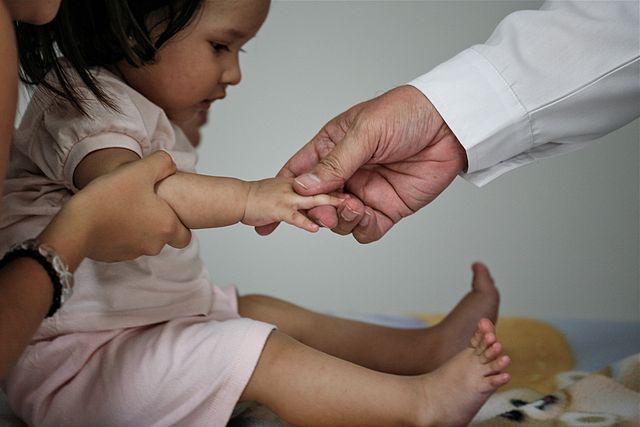 A white toddler with long black hair and dressed in a pick dress, is held by an adult mother. The toddler sits on a table, with their hands extended reaching for another adult's masculine hand in front of the toddler.