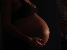 Darkened image of a pregnant woman's torso, showing only a side view of her stomach. She is holding the bottom of her stomach.