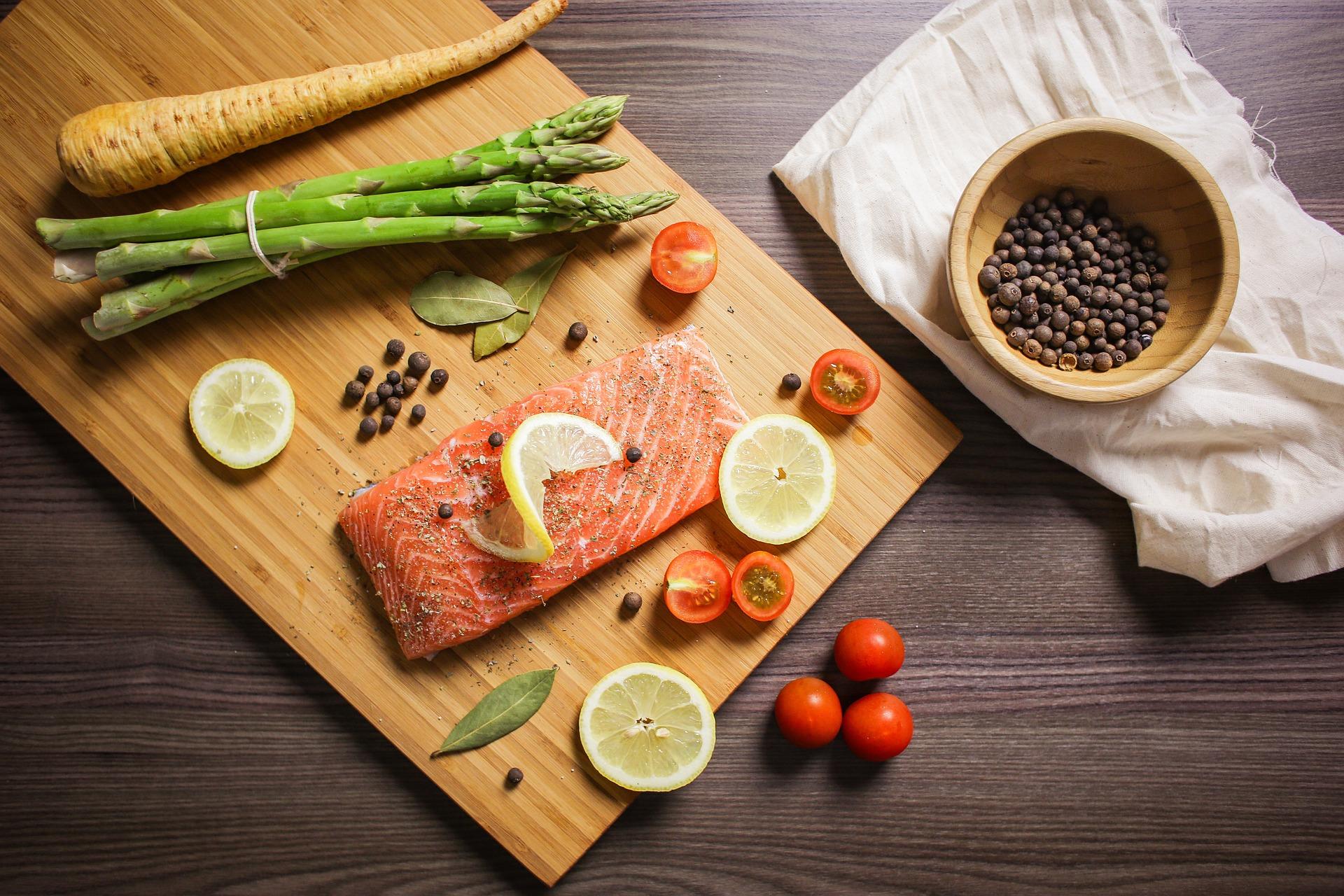 Bird's eye view of a cutting board, displaying vegetables and an uncooked salmon. A bowl of peppercorns are plated to its side.