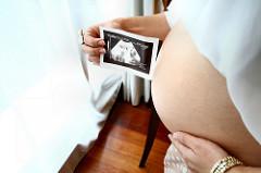 Bird's eye view of the side of a pale skinned pregnant woman, with an exposed stomach, who uses one hand to grasp her stomach, and the other to hold up a sonogram of the baby.