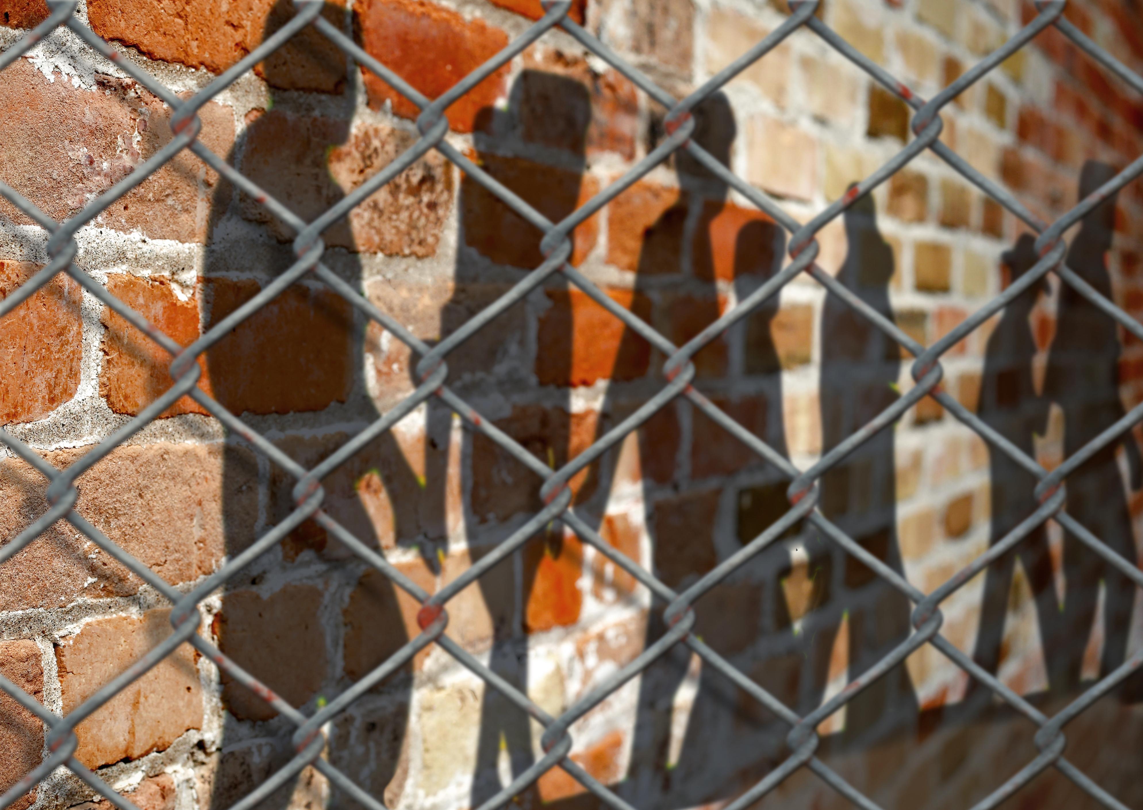 silhouettes of figures cast on a wall behind a cyclone fence. 